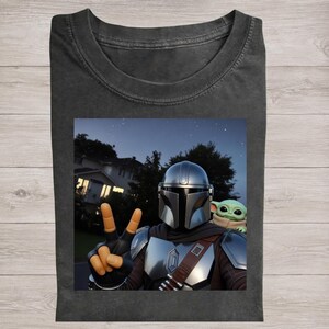 May include: Dark gray t-shirt featuring a graphic of The Mandalorian and Grogu. The Mandalorian is making a peace sign with his fingers. The background shows a night sky with stars and a house.