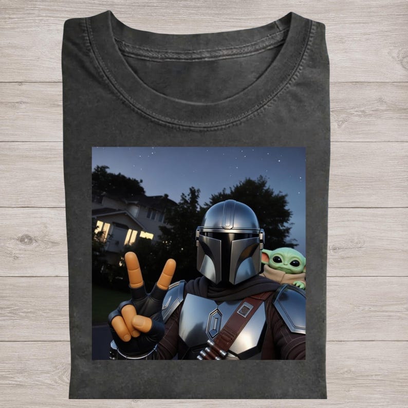 May include: Dark gray t-shirt featuring a graphic of The Mandalorian and Grogu. The Mandalorian is making a peace sign with his fingers. The background shows a night sky with stars and a house.