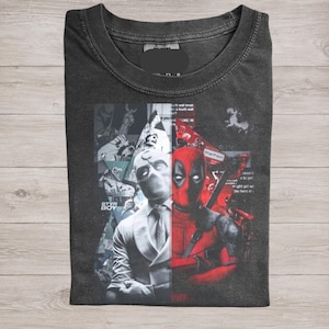 May include: Dark gray t-shirt featuring a split design with Deadpool on the right in red and a character in white on the left. The shirt has a crew neck and a graphic print.