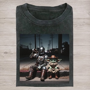 Puede incluir: Camiseta gris oscuro con un gr&aacute;fico de The Mandalorian bebiendo de una taza, sentado junto a Grogu con un sombrero de vaquero. La imagen est&aacute; ambientada en una escena callejera nocturna. La palabra "Trueto" es visible.