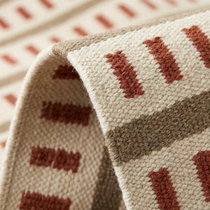Peut inclure: Gros plan sur un tapis tissé beige et marron avec un motif géométrique. Le tapis présente des formes rectangulaires de couleur rouille répétées et une bande marron sur un fond beige. Le bord du tapis est visible, montrant son épaisseur et sa texture.