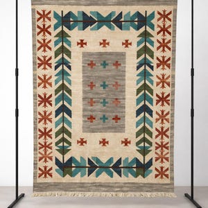 Puede incluir: Alfombra rectangular beige con un patr&oacute;n geom&eacute;trico. La alfombra presenta un rect&aacute;ngulo gris central con cruces rojas, rodeado por un borde de flechas y cruces turquesas, verdes y rojas. La alfombra se exhibe en un soporte de metal negro.