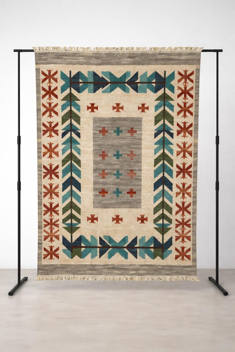 Puede incluir: Alfombra rectangular beige con un patr&oacute;n geom&eacute;trico. La alfombra presenta un rect&aacute;ngulo gris central con cruces rojas, rodeado por un borde de flechas y cruces turquesas, verdes y rojas. La alfombra se exhibe en un soporte de metal negro.