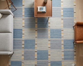 May include: A rectangular area rug featuring a grid pattern of large blue and light gray squares, accented by thin vertical stripes in yellow, white, and dark brown. The rug is woven with a visible texture and lies flat on a wooden floor.