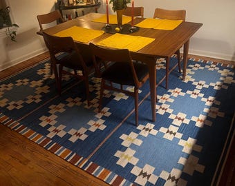 May include: A large rectangular area rug featuring a blue background with a repeating white and brown geometric cross pattern and striped borders, placed under a wooden dining table with four chairs and yellow placemats.