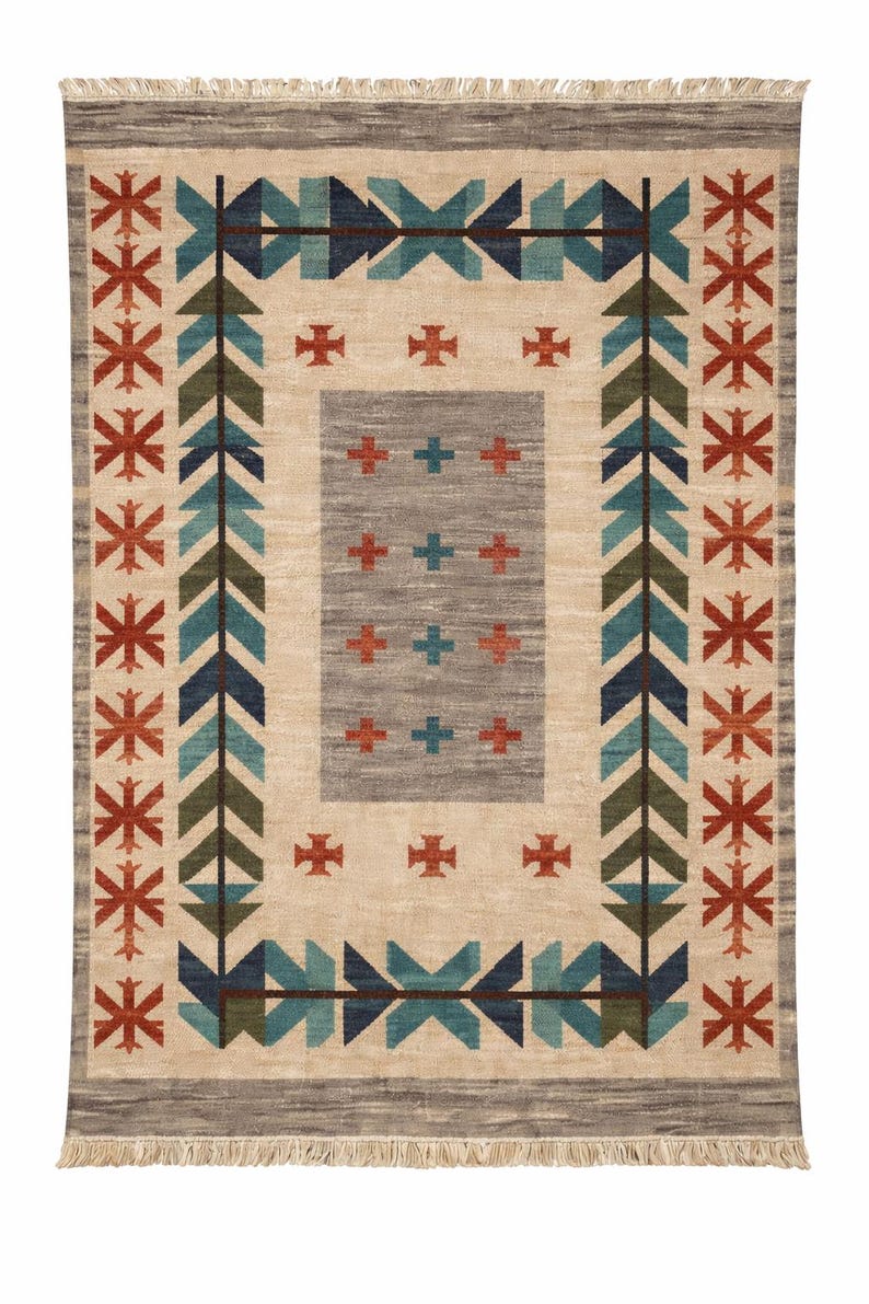 Puede incluir: Alfombra rectangular con un campo central beige y un borde gris. Presenta patrones geom&eacute;tricos en turquesa, verde, azul marino y rojo. El dise&ntilde;o incluye flechas, cruces y motivos de estrellas, creando una est&eacute;tica del suroeste o tribal.