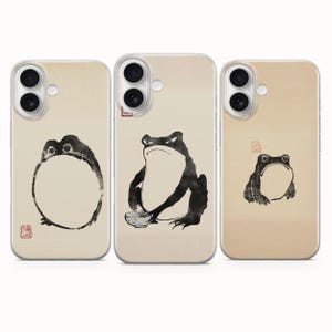 May include: Three phone cases with a beige background and black frog illustrations. The frog designs vary, with one appearing as a circle, another holding a bowl, and the third with a simple outline. Each case has a clear border.