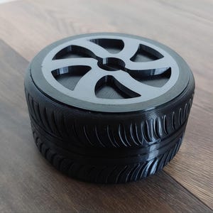 Pode incluir: Um recipiente circular preto em forma de pneu de carro com uma parede lateral com rasto e um design de jante cinzento-escuro na tampa superior. O objeto apresenta um detalhe de cubo central e um acabamento liso, com um corpo cilíndrico e uma tampa.