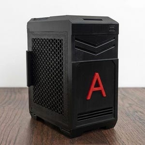 Op de afbeelding: Een zwarte computerkast met een rode "A" op de voorkant. De kast heeft een gaas zijpaneel en schuine lijnen aan de bovenkant. Het staat op een houten oppervlak.