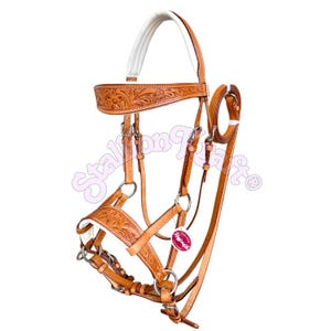 Puede incluir: Una brida de cuero color canela con grabados florales en la frente y la muserola. El cabezal incluye acolchado blanco en la testera y la nariz, herrajes de metal plateado y una pequeña etiqueta con la marca Stallion Craft visible.