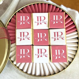 Puede incluir: Una lata redonda dorada llena de bombones. Los bombones están dispuestos en un patrón de cuadrícula, con envoltorios rosas y blancos alternados. Cada envoltorio presenta las iniciales "IR" y las palabras "Romeo & Juliet".