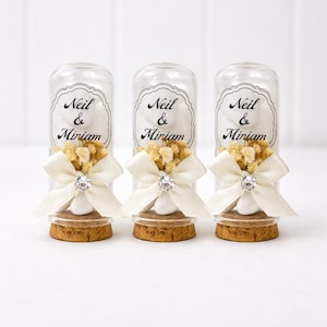 Puede incluir: Tres tubos de vidrio transparente con tapones de corcho, cada uno adornado con un lazo blanco, un pedrería y un pequeño ramo de flores secas. Cada tubo tiene una etiqueta que dice "Neil & Miriam".