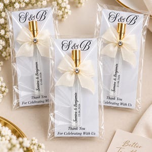 Op de afbeelding: Drie doorzichtige glazen parfumflesjes met gouden doppen, elk versierd met een crèmekleurig lint en een kleine strass-steentje. De flesjes zijn bevestigd op witte kaarten bedrukt met S & B, Semira & Benjamin, 15.06.2026 en Thank You For Celebrating With Us.