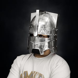 Peut inclure: Un casque de style médiéval de couleur argentée, avec une construction rivetée. Le casque présente un sommet carré et une visière avec des fentes pour les yeux. La personne porte un t-shirt blanc.