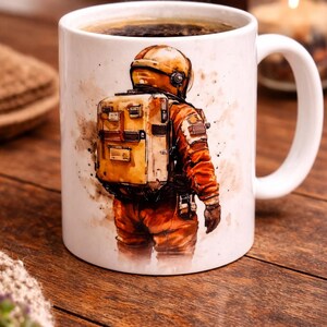 Peut inclure: Mug en céramique blanche avec une illustration à l'aquarelle d'un astronaute en combinaison spatiale orange, avec un sac à dos et un casque jaunes. La tasse est remplie de café noir, et de la vapeur s'en échappe.