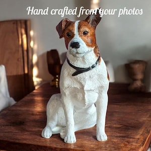 Könnte beinhalten: Eine detaillierte Skulptur eines Jack Russell Terrier-Hundes, überwiegend weiß mit braunen Markierungen an Kopf und Ohren. Der Hund sitzt auf einem Holztisch und trägt ein schwarzes Halsband. Der Text "Hand crafted from your photos" steht über dem Hund.