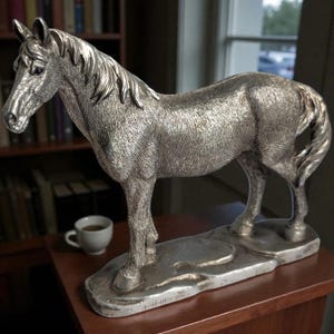 Könnte beinhalten: Eine silberfarbene Pferdestatue, die auf einem rechteckigen Sockel steht. Die Skulptur zeigt die Mähne, den Schweif und die Muskulatur des Pferdes. Eine kleine weiße Tasse steht in der Nähe auf einer Holzoberfläche, mit einem Bücherregal im Hintergrund.