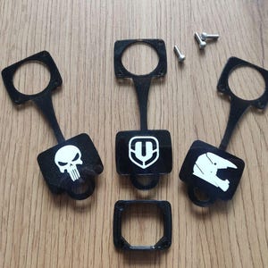 May include: Black plastic keychains with white graphic designs, including a skull, a shield, and a motorcycle helmet. The keychains have a rectangular top with a circular opening and a small loop at the bottom. Small screws are also present.