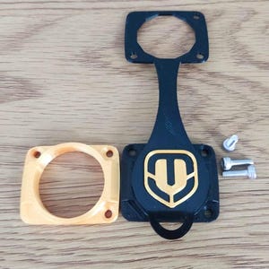Puede incluir: Un conjunto de piezas impresas en 3D, que incluye un emblema negro y dorado, una pieza circular de color naranja claro y varios tornillos. El emblema presenta un diseño dorado sobre un fondo negro. Las piezas están dispuestas sobre una superficie de madera.