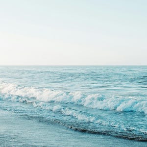 Può includere: Un sereno paesaggio marino con dolci onde che si infrangono sulla riva. L'acqua passa da un turchese chiaro all'orizzonte a un blu più profondo. Il cielo è di un azzurro pallido e morbido, creando un'atmosfera calma.