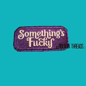 May include: A rectangular embroidered patch with a purple background and a dark purple stitched border. The text "Something's Fucky" is embroidered in a light cream-coloured, stylized cursive font across the centre of the patch.