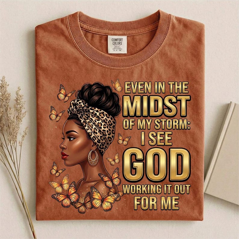 Pu&ograve; includere: T-shirt color ruggine con una grafica di una donna con un copricapo e farfalle. Il testo recita: "Even in the Midst of My Storm: I See God Working It Out For Me."