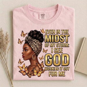 Pu&ograve; includere: T-shirt rosa chiaro con grafica di una donna con un foulard leopardato e farfalle. Il testo recita: "Even in the Midst of My Storm: I See God Working It Out For Me."