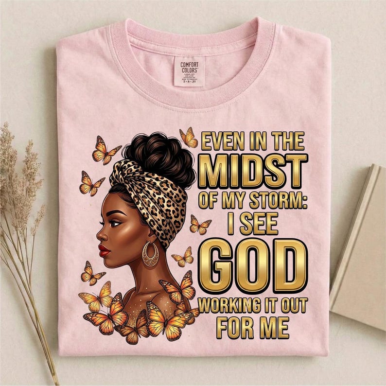 Pu&ograve; includere: T-shirt rosa chiaro con grafica di una donna con un foulard leopardato e farfalle. Il testo recita: "Even in the Midst of My Storm: I See God Working It Out For Me."