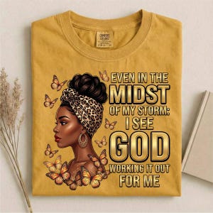 Pu&ograve; includere: T-shirt giallo senape con grafica di una donna con un copricapo leopardato e farfalle. Il testo recita: "Even in the Midst of My Storm: I See God Working It Out For Me."