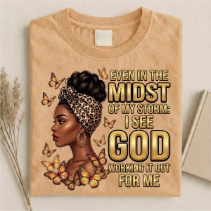 Pu&ograve; includere: T-shirt beige con grafica di una donna con un turbante leopardato e orecchini dorati, circondata da farfalle. Il testo recita: "Anche in mezzo alla mia tempesta: vedo Dio che lo risolve per me."