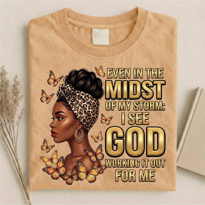 Pu&ograve; includere: T-shirt beige con grafica di una donna con un turbante leopardato e orecchini dorati, circondata da farfalle. Il testo recita: "Anche in mezzo alla mia tempesta: vedo Dio che lo risolve per me."