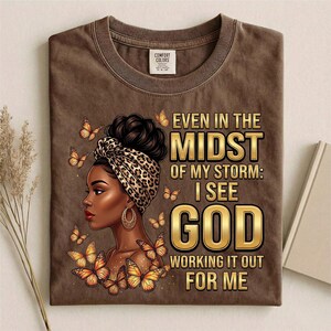 Pu&ograve; includere: T-shirt marrone con grafica di una donna con un copricapo leopardato, orecchini dorati e farfalle. Il testo recita: "Even in the Midst of My Storm: I See God Working It Out For Me."