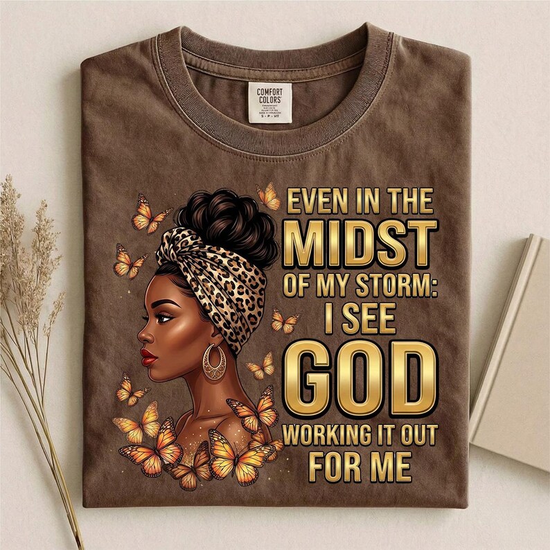 Pu&ograve; includere: T-shirt marrone con grafica di una donna con un copricapo leopardato, orecchini dorati e farfalle. Il testo recita: "Even in the Midst of My Storm: I See God Working It Out For Me."