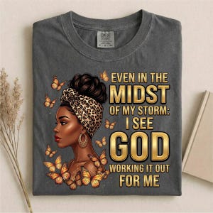 Pu&ograve; includere: T-shirt grigio scuro con una grafica di una donna con un copricapo leopardato e farfalle. Il testo recita: "Even in the Midst of My Storm: I See God Working It Out For Me."