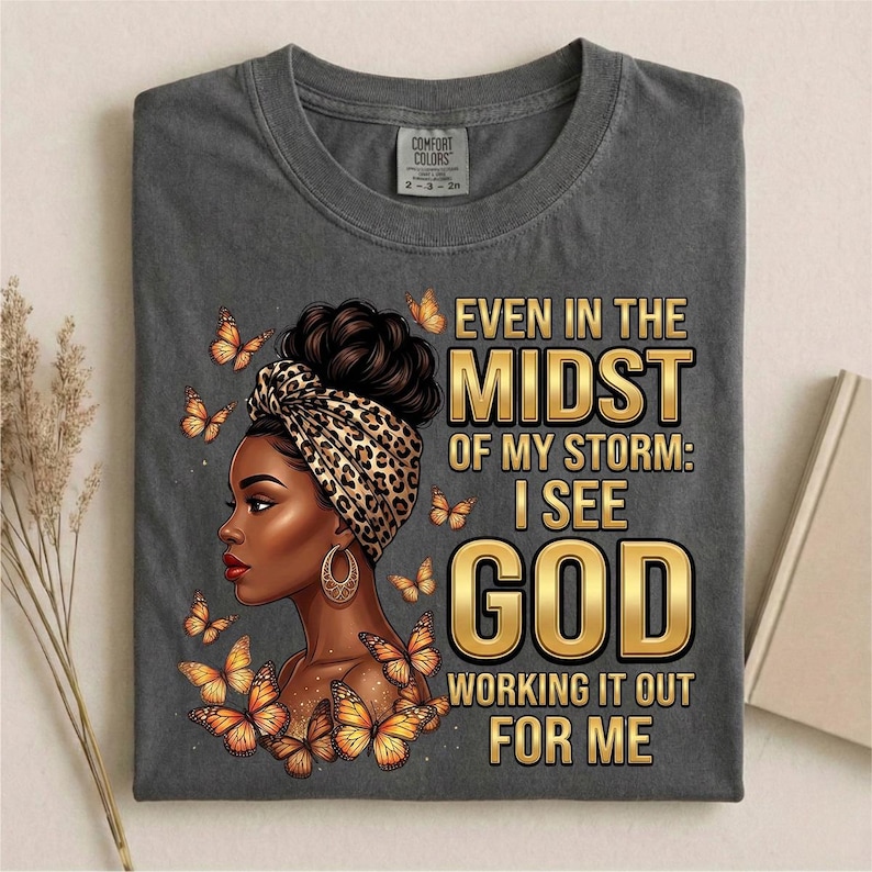 Pu&ograve; includere: T-shirt grigio scuro con una grafica di una donna con un copricapo leopardato e farfalle. Il testo recita: "Even in the Midst of My Storm: I See God Working It Out For Me."