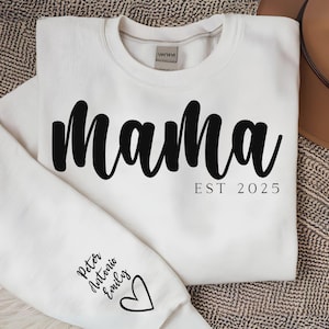 Puede incluir: Sudadera blanca con la palabra "Mama" en letras negras grandes en el pecho. Debajo, "EST 2025". La manga izquierda muestra los nombres "Peter Antonio Emily" y un corazón. Prenda casual y cómoda.
