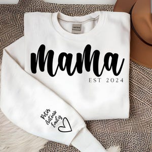 Op de afbeelding: Witte sweatshirt met het woord "Mama" in grote, zwarte letters over de borst, met "EST 2024" eronder. De linkermouw heeft namen en een hartje. Een casual, comfortabel kledingstuk.