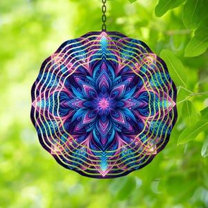 May include: A round, decorative wind spinner with a vibrant, colorful mandala design. The mandala features shades of blue, pink, and purple, with green accents. The spinner hangs from a chain against a blurred green background.