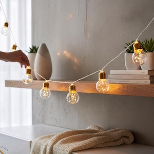 Könnte beinhalten: Lichterkette mit klaren, runden Glühbirnen und warmweißen LED-Leuchten. Die Lichterkette befindet sich auf einem weißen Draht und wird auf einem Holzregal präsentiert. Die Glühbirnen haben einen goldenen Sockel.