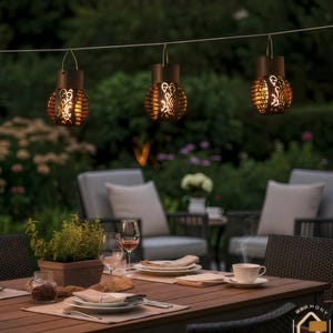Könnte beinhalten: Drei hängende Außenlaternen mit dekorativem Ausschnittdesign, die über einem Holztisch zum Abendessen aufgehängt sind. Die Laternen haben einen warmen, einladenden Schein und schaffen ein gemütliches Ambiente. Die Szene spielt in einem Garten.