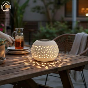 May include: A cream-colored, spherical outdoor solar lantern with leaf-shaped cutouts, casting a warm glow on a wooden table. The lantern has a textured surface and a solar panel on top. The scene includes a pitcher of iced tea and a woven chair.
