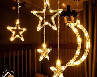 May include: String lights with illuminated stars and a crescent moon. The lights are warm white and hang from a ceiling, creating a decorative ambiance. The lights are strung together, ideal for home decor.
