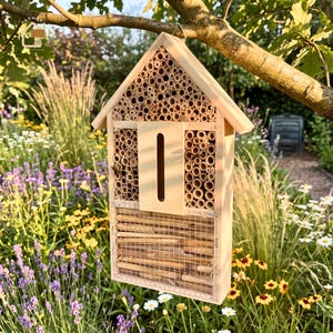 May include: A light-colored wooden insect hotel with a gabled roof, featuring multiple compartments filled with hollow bamboo tubes and stacked twigs. A central vertical slot is present, and the lower section is secured with a metal wire mesh.