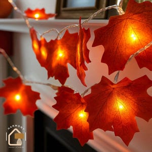 May include: A string of decorative lights with orange maple leaf shapes. Each leaf has a warm, glowing light inside, creating a cosy autumn ambiance. The lights are strung on a thin, silver wire, ideal for seasonal home decoration.