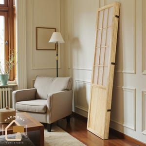 May include: A light-colored wooden room divider with a grid pattern and translucent panels leans against a white wall. The divider is in a room with a beige armchair, a lamp, and a wooden coffee table. The room has a neutral color palette.