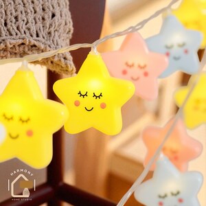 May include: A string of star-shaped fairy lights in yellow, pink, and blue. Each star has a smiling face with closed eyes and rosy cheeks. The lights are strung on a thin white wire, ideal for decorating a child's room or adding a whimsical touch.