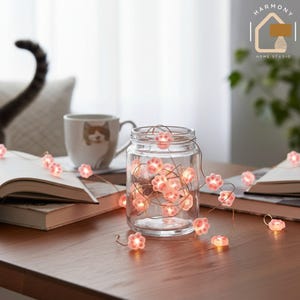 Può includere: Un barattolo di vetro trasparente pieno di luci a forma di zampa di gatto rosa. Il barattolo è su un tavolo di legno, vicino a una tazza con un motivo di gatto e un libro aperto. Le luci sono accese.