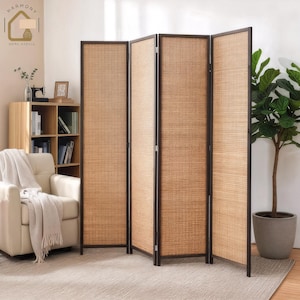 May include: A four-panel room divider with a woven, light brown texture. The frame is dark brown. The divider is standing on a light-colored rug. A potted plant is to the right of the divider. The divider is in a room with a chair and bookshelf.