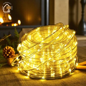 Può includere: Una stringa arrotolata di luci a LED bianco caldo, ideale per scopi decorativi. Le luci sono avvolte in un tubo trasparente e flessibile, creando un bagliore morbido e invitante. Le luci sono su una superficie marrone.