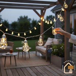 May include: Outdoor string lights with clear, bulb-shaped lights. The lights are strung across a wooden pergola, illuminating a patio setting with seating and small tables. A solar panel is visible, suggesting energy efficiency.